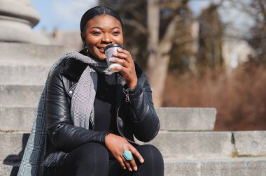 Neşeli, koyu tenli, kahve içmeyi seven, şehir parkında eğlenen, modaya uygun giyinmiş, Afro-Amerikan hippi kız güneşli bir günde dışarıda oturmuş içecek içiyor.