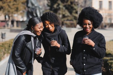 Üç güzel, koyu tenli, uzun saçlı kız bir şehirde duruyor, kahve içiyor ve telefonları kullanıyor.