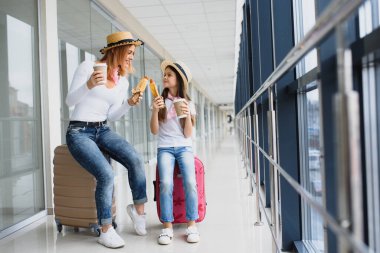 Anne ve kızı havaalanı terminalinde bavullarıyla fast food yiyorlar.