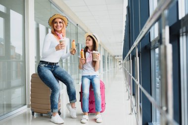 Anne ve kızı havaalanı terminalinde bavullarıyla fast food yiyorlar.