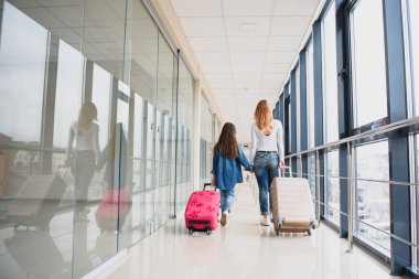Mutlu genç anne ve kızı havaalanı terminalinde bir bavul taşırken yürüyorlar. Yüksek mevsim ve tatil konsepti. Rahatlayın ve yaşam tarzları