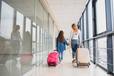 Mutlu genç anne ve kızı havaalanı terminalinde bir bavul taşırken yürüyorlar. Yüksek mevsim ve tatil konsepti. Rahatlayın ve yaşam tarzları