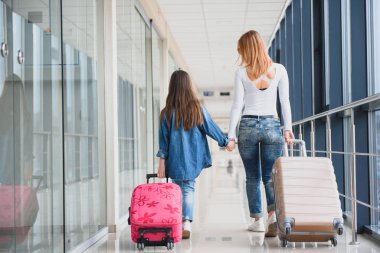 Mutlu genç anne ve kızı havaalanı terminalinde bir bavul taşırken yürüyorlar. Yüksek mevsim ve tatil konsepti. Rahatlayın ve yaşam tarzları
