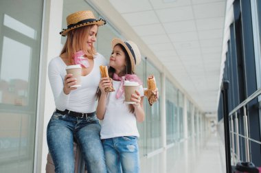 Anne ve kızı bir tatil için havaalanı terminalinde bavullu bir uçak bekliyorlar. Fast food yiyen kızıyla birlikte uçağı bekliyor.
