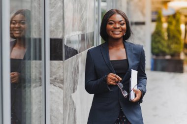 Kendine güvenen iş kadını dışarıda mesaj atıyor. Ofisteki takım elbiseli siyahi kadın cam duvarın yanında duruyor ve cep telefonu kullanıyor. Afrikalı Amerikalı iş kadını konsepti