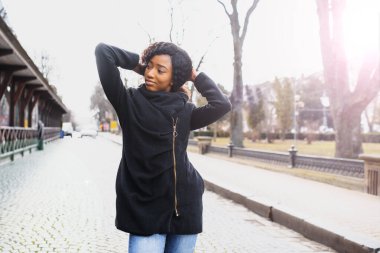 Afro saç stili ve burun halkası olan çekici, mutlu, koyu tenli bir model sabah yürüyüşü sırasında şehir arka planına karşı poz veriyor, beyaz dişlerini gösteren utangaç bir gülümsemeyle aşağıya bakıyor. Parlak güneş