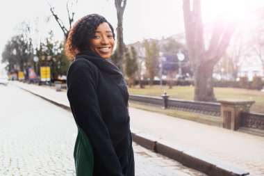 Afro saç stili ve burun halkası olan çekici, mutlu, koyu tenli bir model sabah yürüyüşü sırasında şehir arka planına karşı poz veriyor, beyaz dişlerini gösteren utangaç bir gülümsemeyle aşağıya bakıyor. Parlak güneş