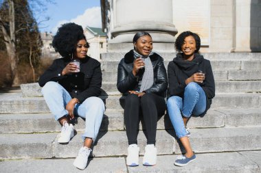 Üç güzel, koyu tenli, uzun saçlı kız bir şehirde duruyor, kahve içiyor ve telefonları kullanıyor.