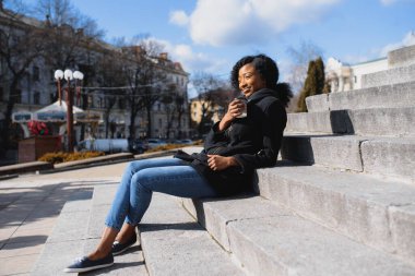 Neşeli, koyu tenli, kahve içmeyi seven, şehir parkında eğlenen, modaya uygun giyinmiş, Afro-Amerikan hippi kız güneşli bir günde dışarıda oturmuş içecek içiyor.
