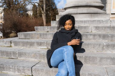 Düşünceli, gözlüklü, kahve içen, sokak merdivenlerinde oturan Afrikalı bir kadın.