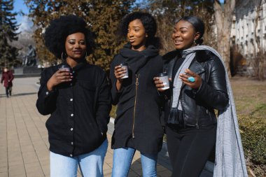 Üç güzel, koyu tenli, uzun saçlı kız bir şehirde duruyor, kahve içiyor ve telefonları kullanıyor.