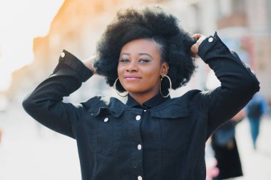 Afro saç stili ve burun halkası olan çekici, mutlu, koyu tenli bir model sabah yürüyüşü sırasında şehir arka planına karşı poz veriyor, beyaz dişlerini gösteren utangaç bir gülümsemeyle aşağıya bakıyor. Parlak güneş