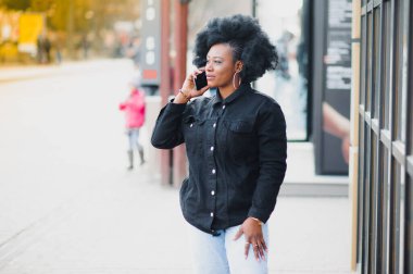 Afro saç stili ve burun halkası olan çekici, mutlu, koyu tenli bir model sabah yürüyüşü sırasında şehir arka planına karşı poz veriyor, beyaz dişlerini gösteren utangaç bir gülümsemeyle aşağıya bakıyor. Parlak güneş