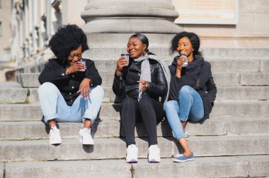 Üç güzel, koyu tenli, uzun saçlı kız bir şehirde duruyor, kahve içiyor ve telefonları kullanıyor.