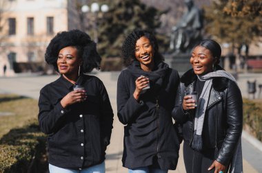 Üç güzel, koyu tenli, uzun saçlı kız bir şehirde duruyor, kahve içiyor ve telefonları kullanıyor.