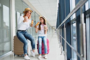 Anne ve kızı havaalanı terminalinde bavullarıyla fast food yiyorlar.