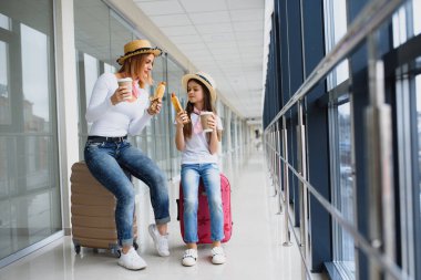 Anne ve kızı havaalanı terminalinde bavullarıyla fast food yiyorlar.