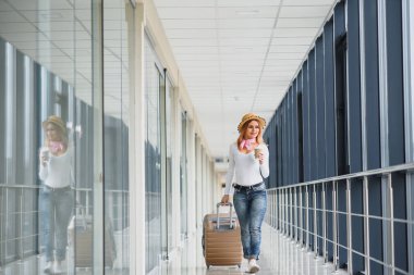 Gündelik giysiler giyen genç bir kadın havaalanında poz veriyor, çanta tutuyor ve kalkış pistinde bagajın yanında bekliyor..