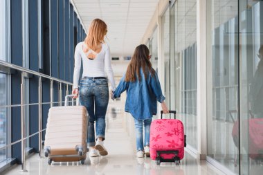 Mutlu genç anne ve kızı havaalanı terminalinde bir bavul taşırken yürüyorlar. Yüksek mevsim ve tatil konsepti. Rahatlayın ve yaşam tarzları