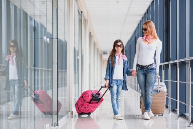 Mutlu genç anne ve kızı havaalanı terminalinde bir bavul taşırken yürüyorlar. Yüksek mevsim ve tatil konsepti. Rahatlayın ve yaşam tarzları