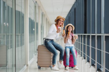Anne ve kızı bir tatil için havaalanı terminalinde bavullu bir uçak bekliyorlar. Fast food yiyen kızıyla birlikte uçağı bekliyor.