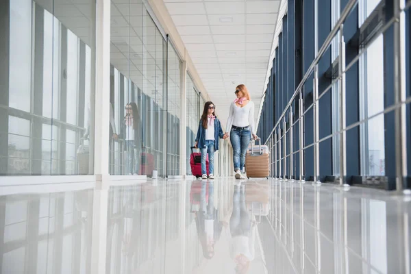 Beautiful mother and her stylish cute daughter walk together and pull a pink suitcase at airport. High season and vacation concept. Relax and lifestyles