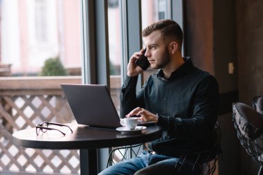 Kafede laptopla çalışırken cep telefonuyla konuşan genç bir işadamı.