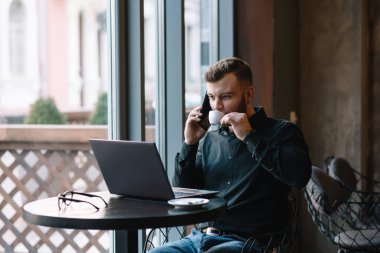 Kafede laptopla çalışırken cep telefonuyla konuşan genç bir işadamı.