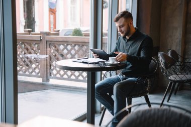 Yakışıklı iş adamı dijital tablet kullanıyor ve şehir merkezindeki kafede otururken kahve içiyor.
