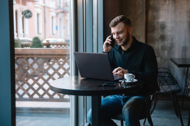 Kafede laptopla çalışırken cep telefonuyla konuşan genç bir işadamı.