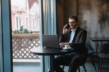 Kafede laptopla çalışırken cep telefonuyla konuşan genç bir işadamı.