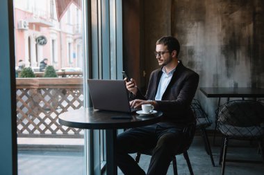 Kafede laptopla çalışırken cep telefonuyla konuşan genç bir işadamı.