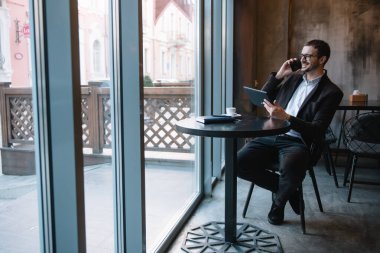 Kafede tablet üzerinde çalışırken cep telefonuyla konuşan genç işadamı