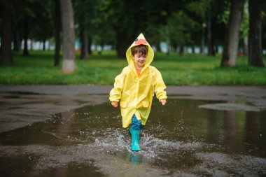 Küçük çocuk yağmurlu yaz parkında oynuyor. Şemsiyeli çocuk, su geçirmez palto ve botlar yağmurda çamurlu su birikintisine atlıyor. Yazın yağmurda yürüyen çocuk her türlü hava koşulunda dışarıda eğleniyor. Mutlu çocukluk