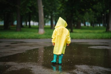 Küçük çocuk yağmurlu yaz parkında oynuyor. Şemsiyeli çocuk, su geçirmez palto ve botlar yağmurda çamurlu su birikintisine atlıyor. Yazın yağmurda yürüyen çocuk her türlü hava koşulunda dışarıda eğleniyor. Mutlu çocukluk