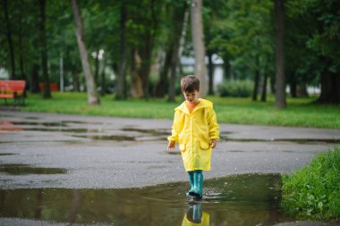 Küçük çocuk yağmurlu yaz parkında oynuyor. Şemsiyeli çocuk, su geçirmez palto ve botlar yağmurda çamurlu su birikintisine atlıyor. Yazın yağmurda yürüyen çocuk her türlü hava koşulunda dışarıda eğleniyor. Mutlu çocukluk