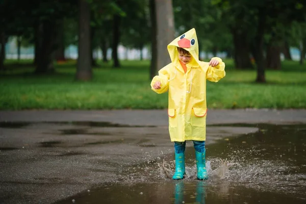 雨の夏の公園で遊んでいる男の子 雨の中で水たまりや泥の中でジャンプ傘 防水コートやブーツと子供 子供は夏の雨の中を歩くどんな天気でも屋外の楽しみ 幸せな子供時代 — ストック写真