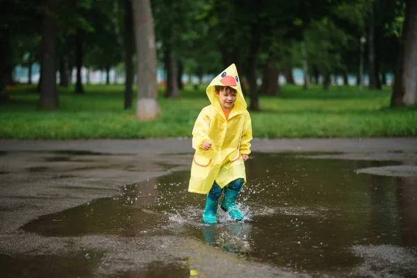 雨の夏の公園で遊んでいる男の子 雨の中で水たまりや泥の中でジャンプ傘 防水コートやブーツと子供 子供は夏の雨の中を歩くどんな天気でも屋外の楽しみ 幸せな子供時代 — ストック写真