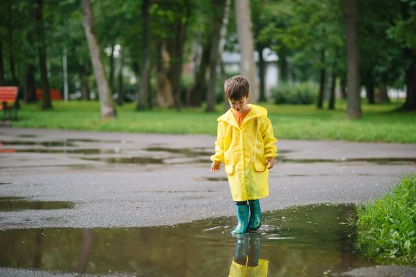 Little boy playing in rainy summer park. Child with umbrella, waterproof coat and boots jumping in puddle and mud in the rain. Kid walking in summer rain Outdoor fun by any weather. happy childhood