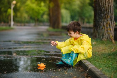 Çocuk su birikintisinde oyuncak tekneyle oynuyor. Çocuk yağmur altında dışarıda oynuyor. Genç çocuklar için sonbahar yağmurlu hava aktivitesi. Çamurlu su birikintilerinde atlayan çocuk. Bebek için su geçirmez ceket ve botlar. Yaz tatili. Mutlu çocukluk