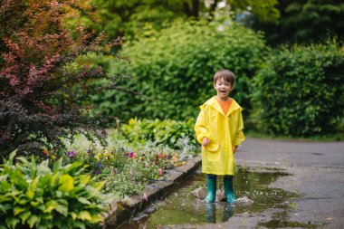 Küçük çocuk yağmurlu yaz parkında oynuyor. Şemsiyeli çocuk, su geçirmez palto ve botlar yağmurda çamurlu su birikintisine atlıyor. Yazın yağmurda yürüyen çocuk her türlü hava koşulunda dışarıda eğleniyor. Mutlu çocukluk