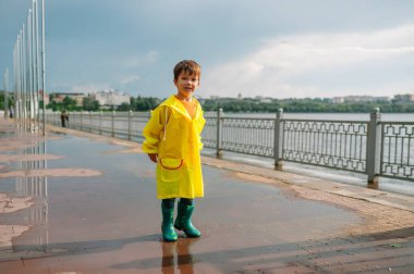 Küçük çocuk yağmurlu yaz parkında oynuyor. Şemsiyeli çocuk, su geçirmez palto ve botlar yağmurda çamurlu su birikintisine atlıyor. Yazın yağmurda yürüyen çocuk her türlü hava koşulunda dışarıda eğleniyor. Mutlu çocukluk