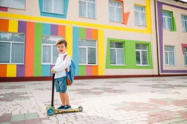 Tekme scooter 'ı olan genç bir çocuk, renkli duvarın yanında. Gözlüklü mutlu çocuk ilk kez okula gidiyor. Sırt çantası ve kitabı olan bir çocuk. Dersler başlıyor. Sonbaharın ilk günü.