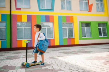 Tekme scooter 'ı olan genç bir çocuk, renkli duvarın yanında. Gözlüklü mutlu çocuk ilk kez okula gidiyor. Sırt çantası ve kitabı olan bir çocuk. Dersler başlıyor. Sonbaharın ilk günü.