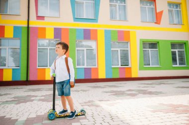 Tekme scooter 'ı olan genç bir çocuk, renkli duvarın yanında. Gözlüklü mutlu çocuk ilk kez okula gidiyor. Sırt çantası ve kitabı olan bir çocuk. Dersler başlıyor. Sonbaharın ilk günü.