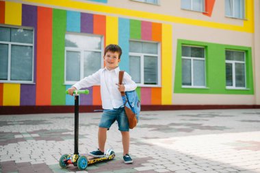 Tekme scooter 'ı olan genç bir çocuk, renkli duvarın yanında. Gözlüklü mutlu çocuk ilk kez okula gidiyor. Sırt çantası ve kitabı olan bir çocuk. Dersler başlıyor. Sonbaharın ilk günü.