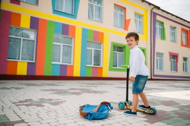 Tekme scooter 'ı olan genç bir çocuk, renkli duvarın yanında. Gözlüklü mutlu çocuk ilk kez okula gidiyor. Sırt çantası ve kitabı olan bir çocuk. Dersler başlıyor. Sonbaharın ilk günü.