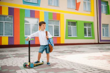 Tekme scooter 'ı olan genç bir çocuk, renkli duvarın yanında. Gözlüklü mutlu çocuk ilk kez okula gidiyor. Sırt çantası ve kitabı olan bir çocuk. Dersler başlıyor. Sonbaharın ilk günü.