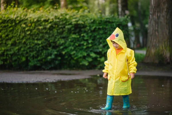 雨の夏の公園で遊んでいる男の子 雨の中で水たまりや泥の中でジャンプ傘 防水コートやブーツと子供 子供は夏の雨の中を歩くどんな天気でも屋外の楽しみ 幸せな子供時代 — ストック写真