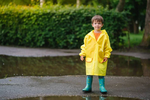 雨の夏の公園で遊んでいる男の子 雨の中で水たまりや泥の中でジャンプ傘 防水コートやブーツと子供 子供は夏の雨の中を歩くどんな天気でも屋外の楽しみ 幸せな子供時代 — ストック写真